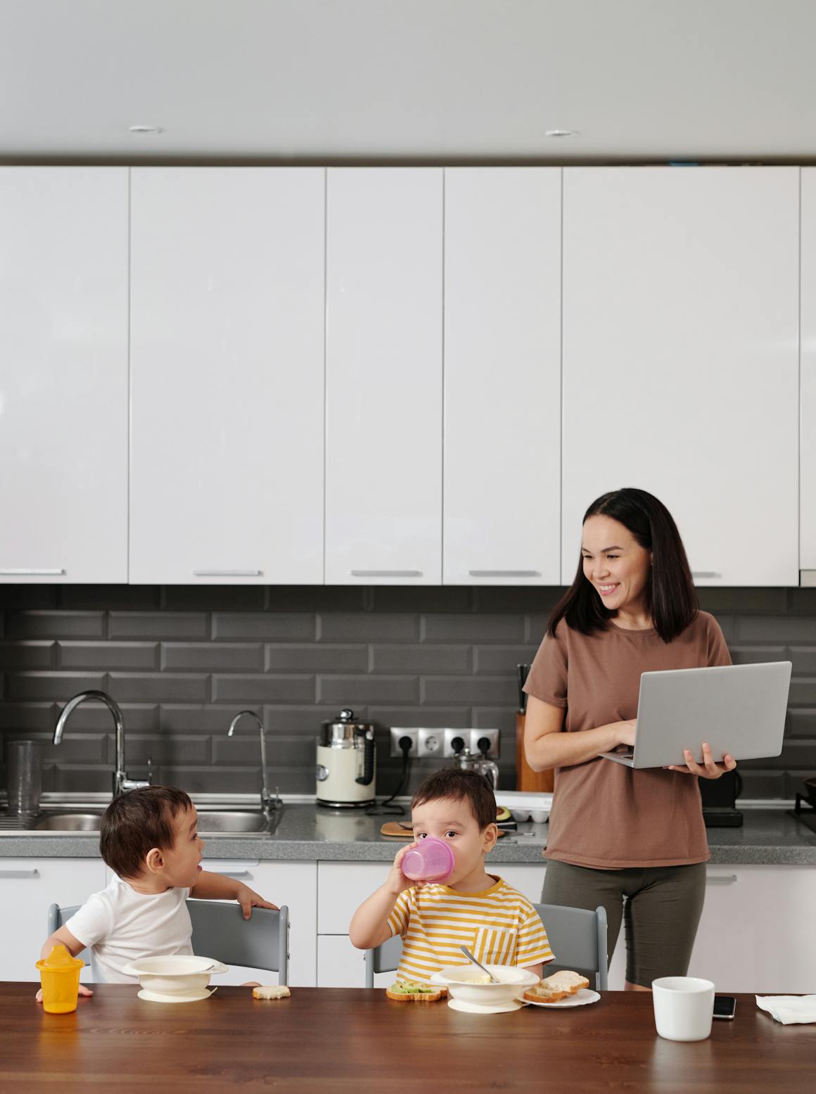 A mother balances work on her laptop while supervising her children during breakfast in a modern kitchen.