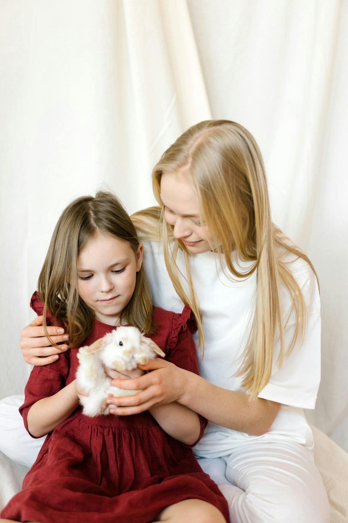 Two sisters smiling and cuddling a white rabbit indoors, promoting togetherness.