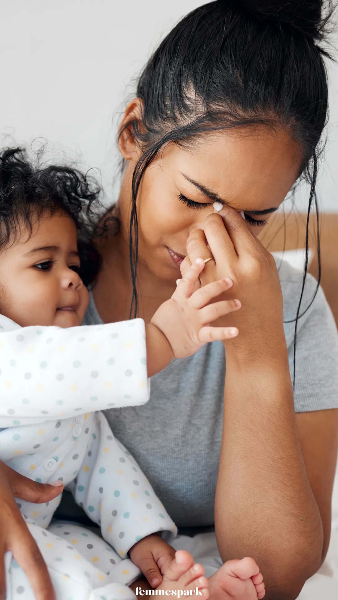 A Black mother looking both tired and loving while her baby holds onto her, depicting the emotional rollercoaster and sleep deprivation common in postpartum recovery.

